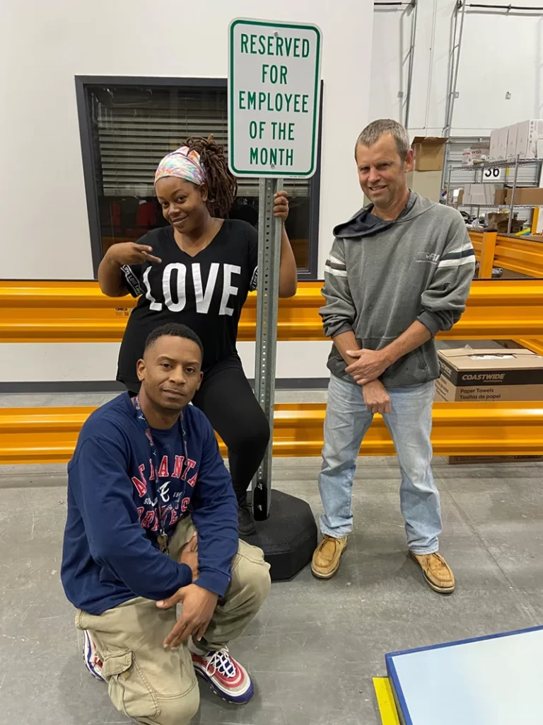 Three people pose indoors near a "Reserved for Employee of the Month" sign in the warehouse. One person is leaning on the sign, while the other two crouch beside it, highlighting the teamwork that keeps our logistics running smoothly.