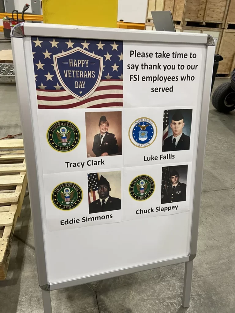 A standing sign displays "Happy Veterans Day" at the top, with a request to thank four FSI employees who served: Tracy Clark, Luke Fallis, Eddie Simmons, Chuck Slappey—each listed with their photos and branch seals—highlighting their vital roles in logistics and warehouse operations.