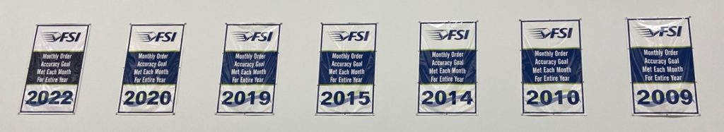 Banners display "Monthly Order Accuracy Goal Met Each Month for Entire Year" with years 2022, 2019, 2015, 2014, 2010, and 2009 under the FSI logo in the fulfillment warehouse.