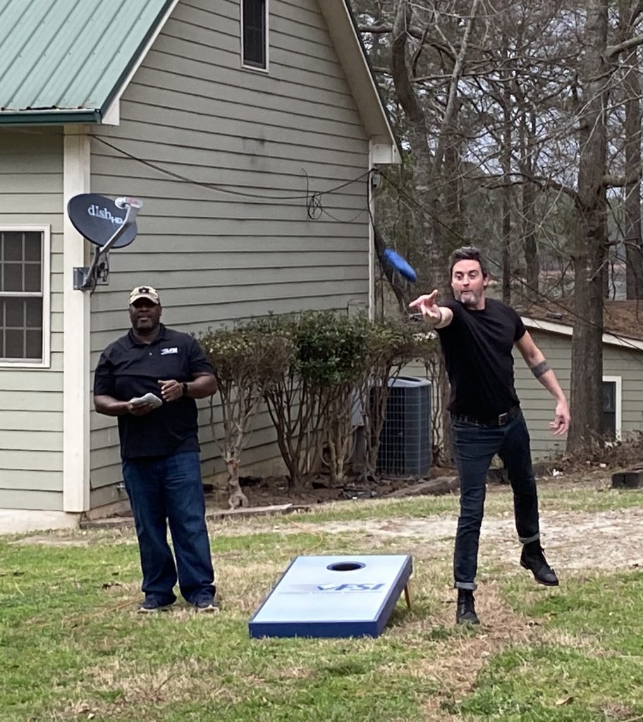Two people are playing cornhole in a yard. One is tossing a bag while the other stands by, perhaps perfecting their shipping technique. A house with a satellite dish is in the background.