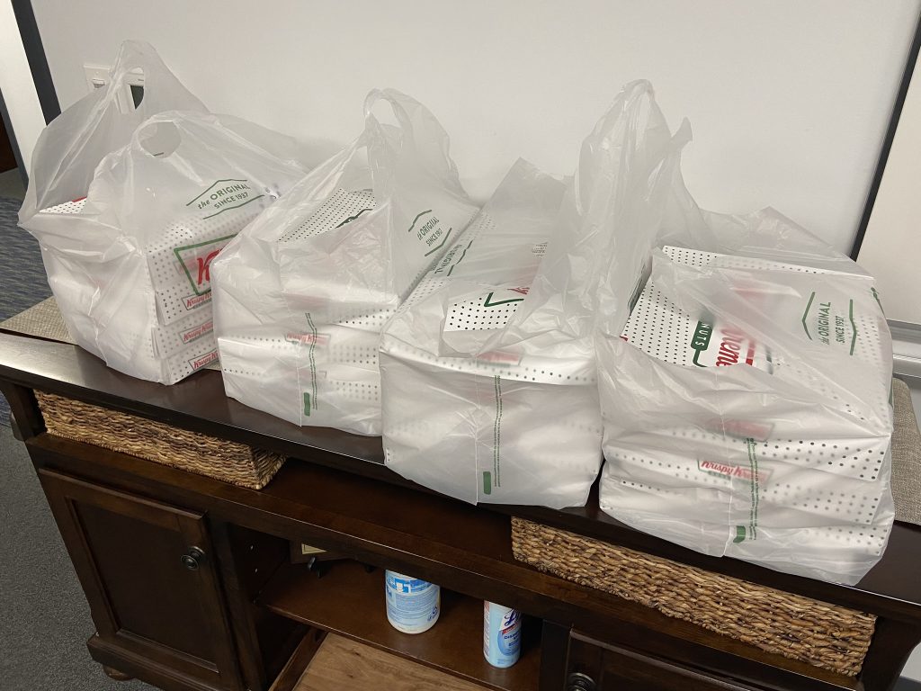 Four plastic bags containing boxes of doughnuts are placed on a wooden side table, with cleaning supplies visible on the lower shelf, indicating the fulfillment of a sweet craving and readiness for quick cleanup.