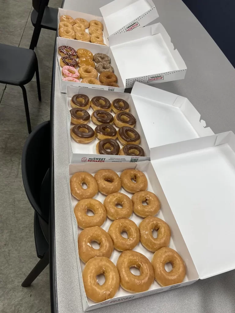 Boxes of assorted donuts on a table, mostly glazed with some chocolate-covered and sprinkled options. Empty chairs visible around the table, as if someone had just finished picking their favorite treats.