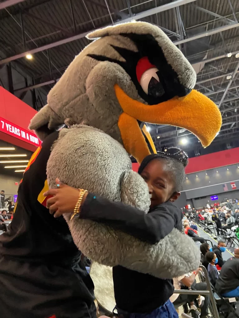 A child hugging a large, gray bird mascot in an indoor sports arena, with spectators and stacks of boxes in the background.