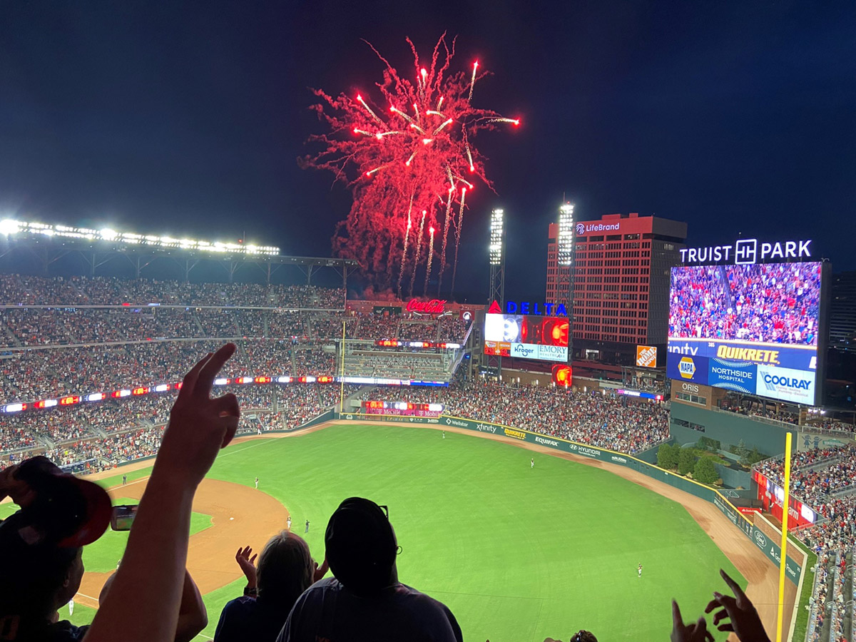 Fireworks burst over a packed baseball stadium at night, as seen from the stands with fans raising their hands in celebration. Amidst the cheers, the logistics of such a grand event are evident as the scoreboard and field come into view.