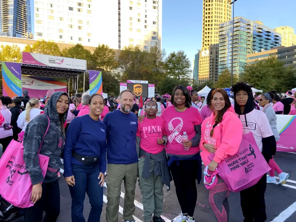 A group of people stands together outdoors, participating in a fundraising event. They are wearing pink and holding bags with event-related text, reminiscent of careful logistics planning. A stage and tall buildings are visible in the background.