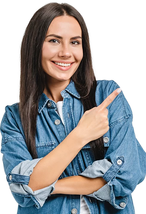 A smiling woman with long dark hair, wearing a denim shirt, points to her right with one hand against a plain background, exuding a sense of fulfillment.