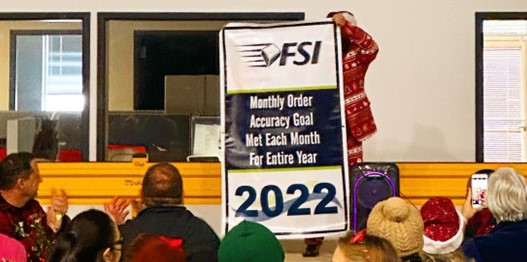 A person in a festive outfit reveals a banner that reads, "FSI Monthly Order Accuracy Goal Met Each Month For Entire Year 2022," while people in the warehouse applaud.