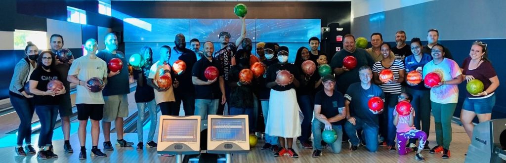A group of people pose with bowling balls at a bowling alley, standing in front of lanes and score monitors, resembling a team ready for action in a bustling logistics warehouse.