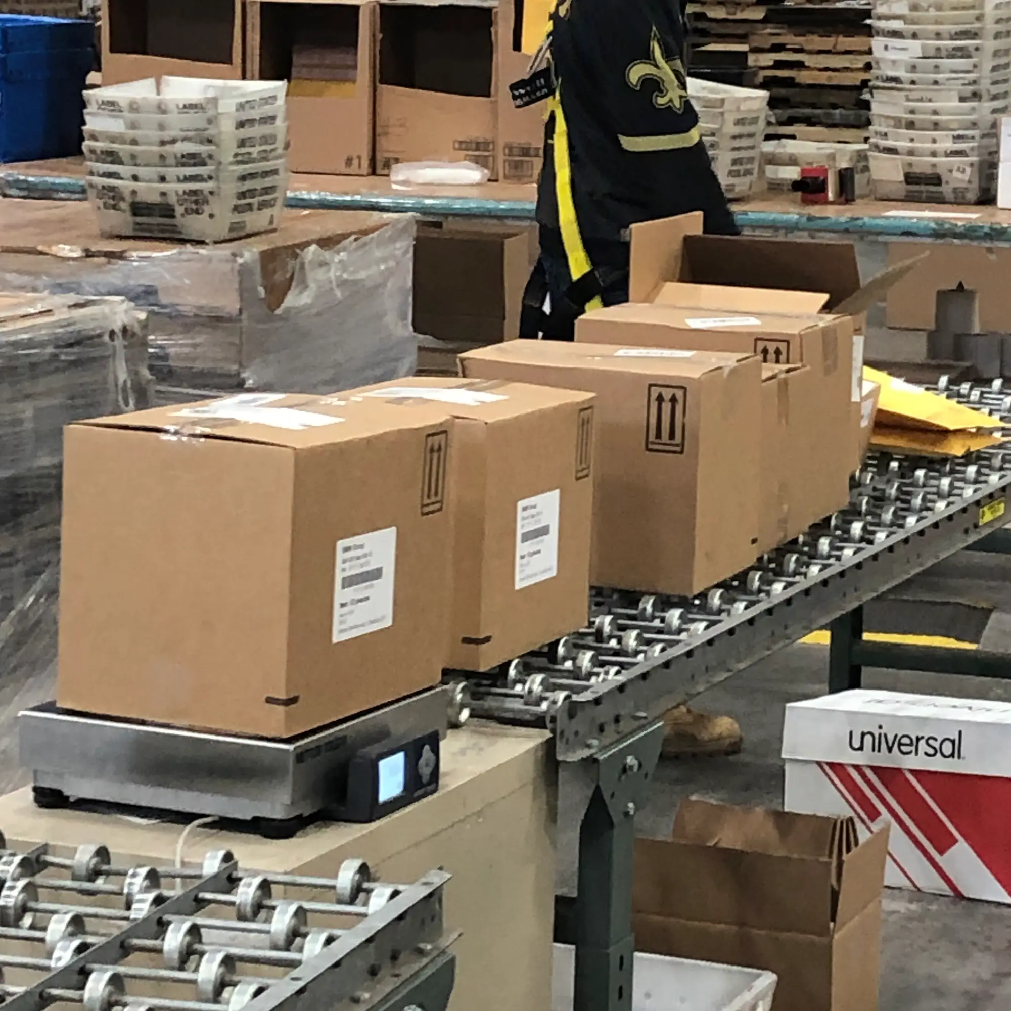 Boxes on a conveyor belt in a warehouse, with an employee in the background wearing a black and yellow shirt, diligently picking items to streamline the fulfillment process.