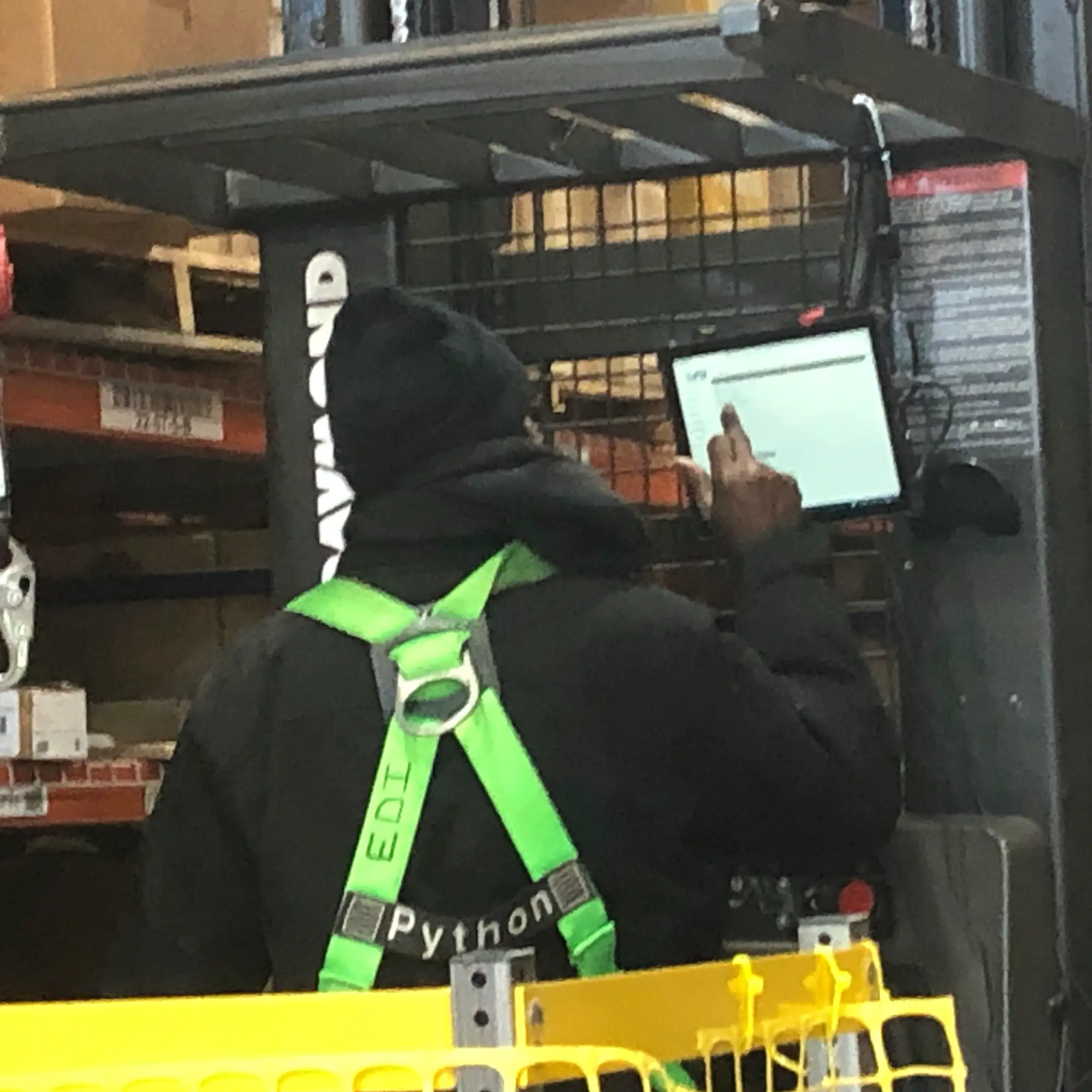 A person wearing a bright green safety harness operates a forklift equipped with a touchscreen interface in the bustling fulfillment warehouse.
