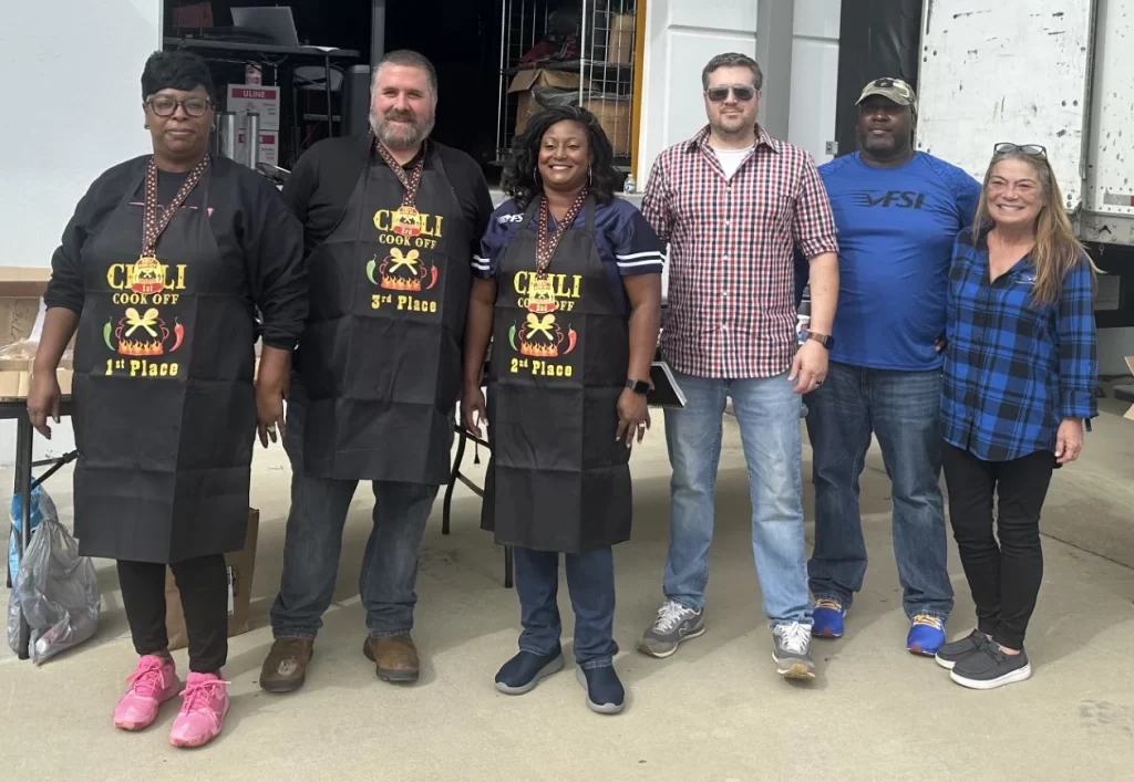 Six people, including two wearing "Chili Cook Off 3rd Place" aprons, stand side by side outdoors, smiling at the camera while holding boxes.