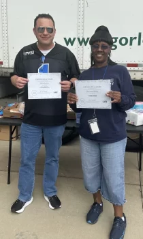 Two individuals stand side by side holding certificates of award. They are outside in front of a shipping truck, wearing casual clothing and smiling at the camera.