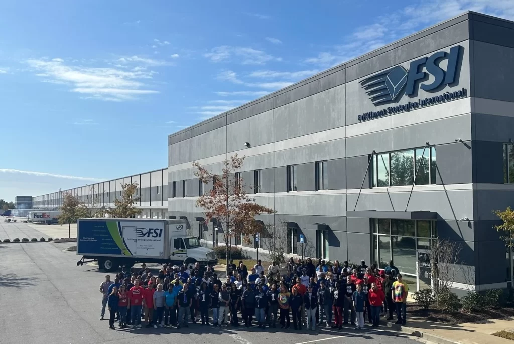 A large group of people are gathered outside a grey building with the logo "FSI" (Fulfillment Strategies International) on it. There is a truck with matching branding parked nearby, hinting at the bustling logistics and picking activities within the warehouse.
