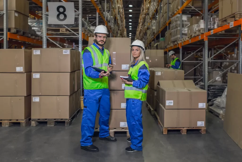 Two warehouse workers in safety gear and hard hats holding tablets, discussing picking and shipping processes in front of stacked cardboard boxes in a large warehouse aisle. Another worker is visible in the background, busy with logistics tasks.