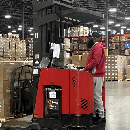 A man in a red jacket operates a forklift in a warehouse, surrounded by stacked boxes and shelves filled with shipping inventory.