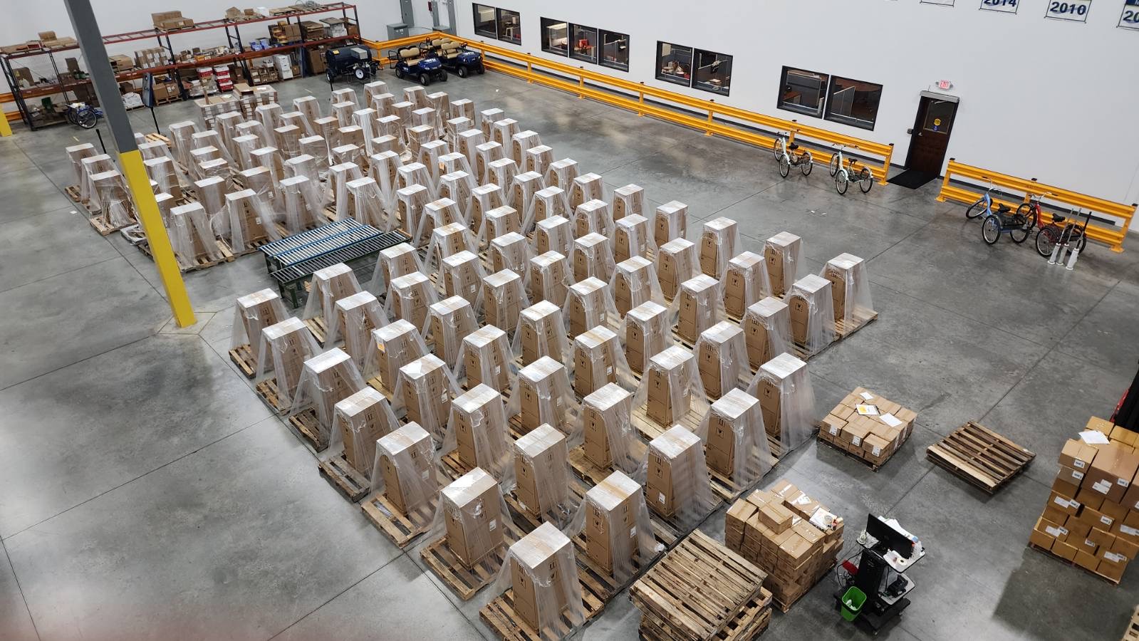 A warehouse with numerous stacked pallets containing boxed items, orderly arranged in rows. Several bicycles are parked along a wall, reflecting the bustling logistics environment, with shelves holding various items nearby for easy picking.