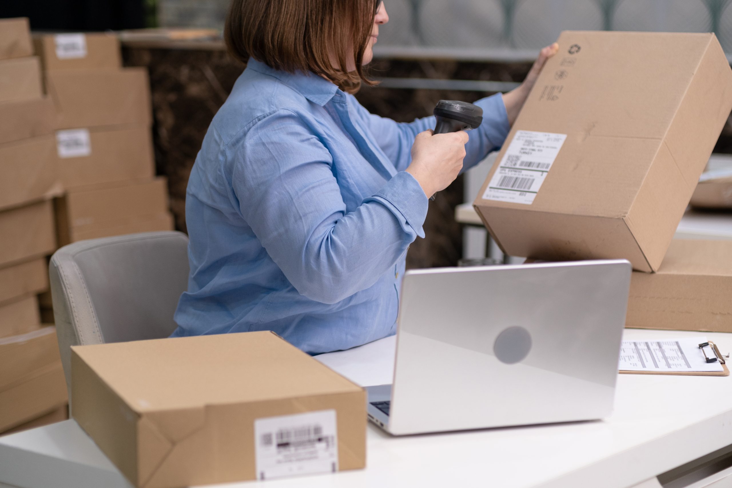 Person in a blue shirt scanning a cardboard box near a laptop and other boxes on a table in a warehouse-like shipping setting.