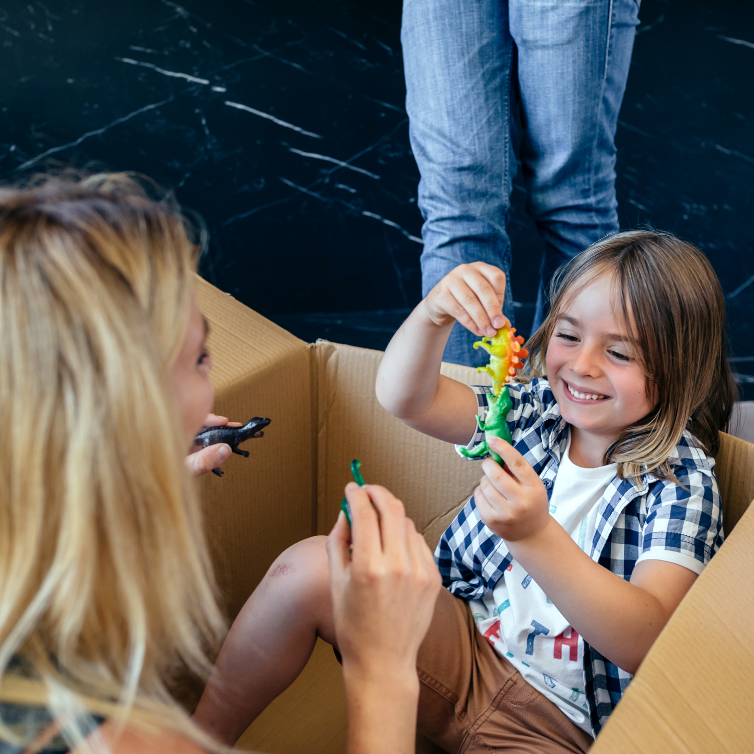 A child sits in a cardboard box holding a toy, smiling at an adult who is also holding a toy, as if they were playing in a busy warehouse. Another person is standing nearby, adding to the joy of this imaginative scene.