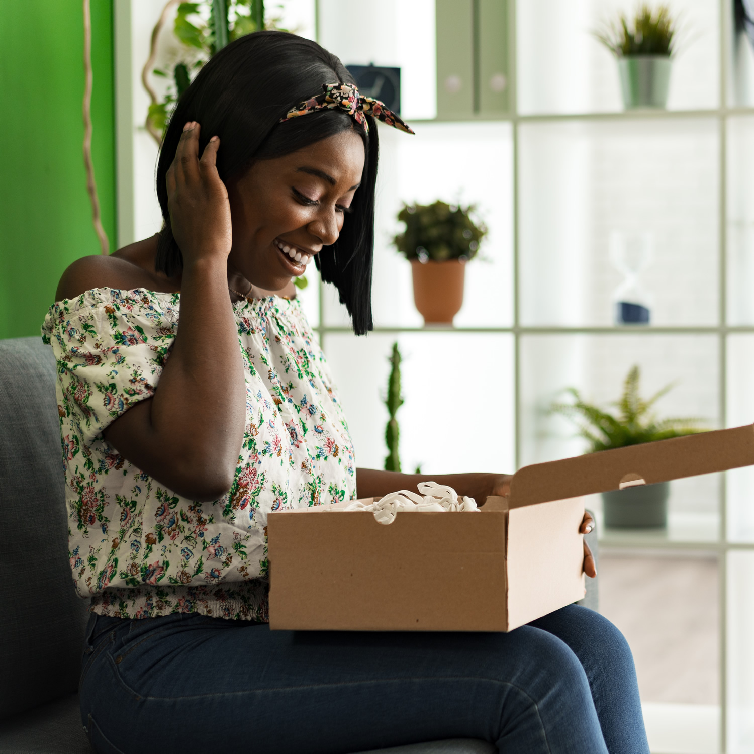 A woman wearing a floral top and headband sits on a couch, smiling while opening a cardboard box. Surrounded by plants and shelves, the scene perfectly captures the joy of home deliveries straight from the warehouse.