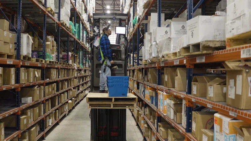 A worker in a warehouse stands on a forklift platform, sorting items on high shelves, surrounded by aisles of stacked boxes, ensuring smooth logistics and efficient fulfillment.