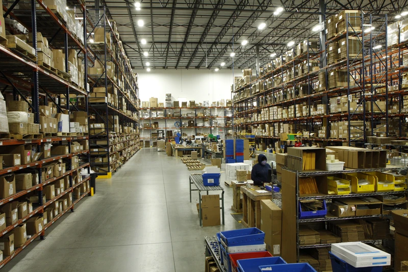 A large, organized warehouse with high shelves filled with boxed inventory. Workers are seen sorting, picking, and packing items at workstations. The aisle is wide and clear, ensuring smooth logistics for efficient shipping.