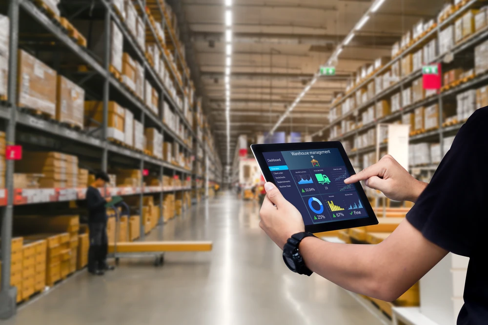 A person using a tablet to manage inventory in a large warehouse with high shelves stacked with various boxes, ensuring efficient picking and fulfillment processes.