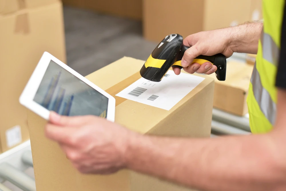 A person in a high-visibility vest scans a barcode on one of the shipping boxes with a handheld scanner, while holding a tablet, ensuring smooth fulfillment operations.