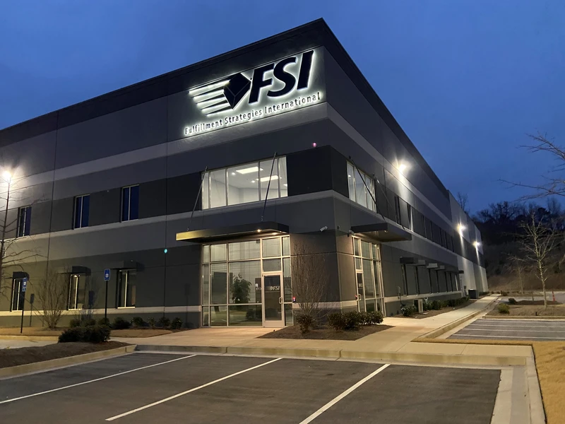 Exterior view of a large warehouse at dusk with the sign "FSI Fulfillment Strategies International" above the entrance. The building has several windows, and the parking lot, usually bustling with shipping trucks and boxes, is empty.