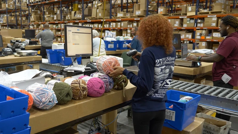 Employees bustling in a logistics warehouse, where a woman is scanning balls of yarn in front of a computer while others meticulously organize products and boxes on shelves and tables.