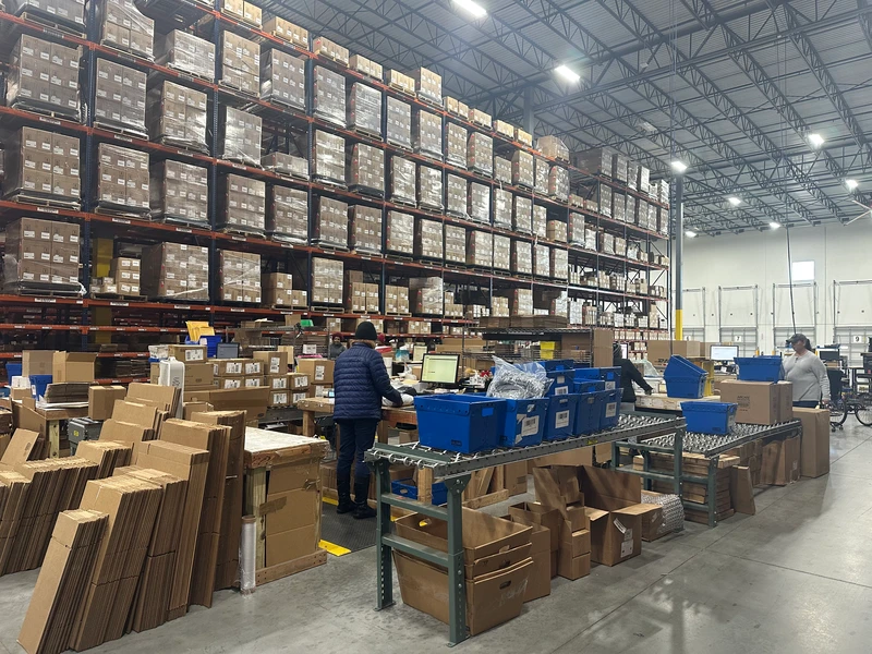 Warehouse scene with workers picking and organizing packages. Shelves stacked with boxes reach the ceiling. Workstations filled with packing materials and blue bins. Elevated view of high, lit ceiling.