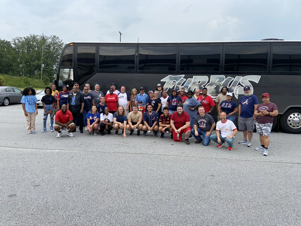 A group of people pose for a photo in front of a large black bus in a parking lot on a cloudy day, surrounded by boxes ready for shipping.