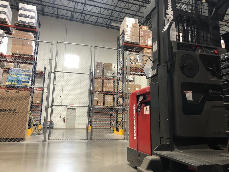 A warehouse interior showing a forklift next to shelves stocked with shipping boxes. A fenced security gate is visible in the background.