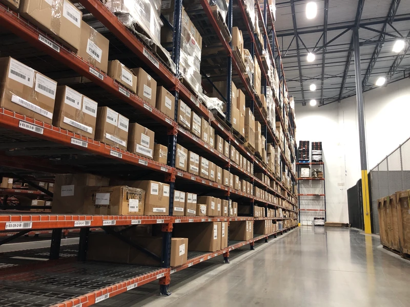 Warehouse interior with tall metal shelves filled with stacked cardboard boxes. The floor is made of polished concrete, perfect for the efficient picking process. The ceiling features metal beams and bright lighting, all seamlessly integrated into a high-functioning logistics environment.