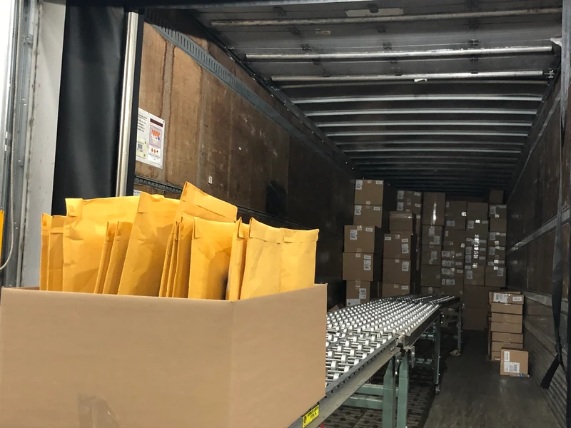 A large cardboard box containing yellow padded envelopes sits on a conveyor belt inside a truck trailer partially filled with brown cardboard boxes, ready for shipping and fulfillment.