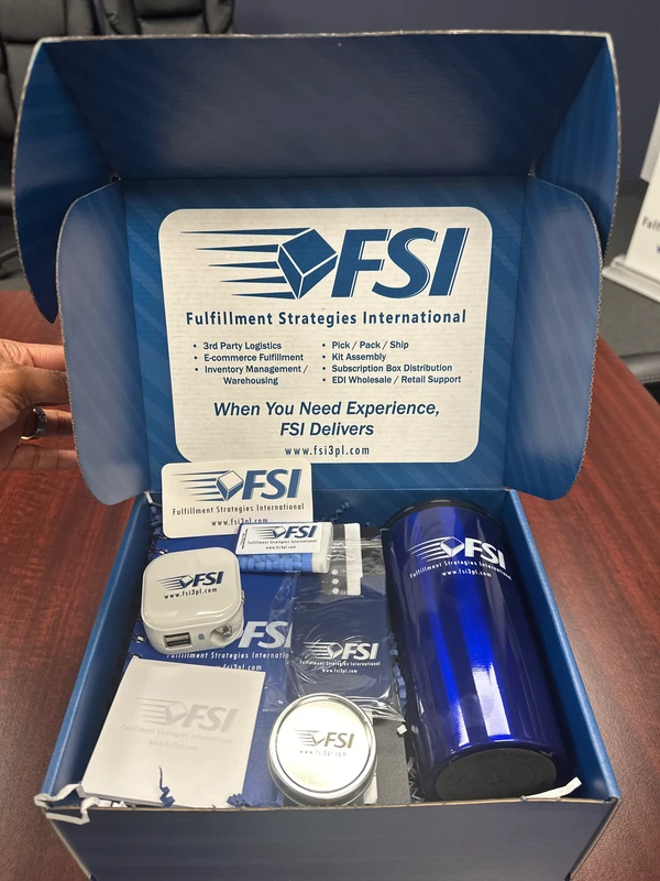 A blue box containing marketing materials for Fulfillment Strategies International, including a tumbler, mints, pen, notebook, and branded pamphlets, displayed on a wooden table—perfectly organized to highlight the importance of efficient logistics.
