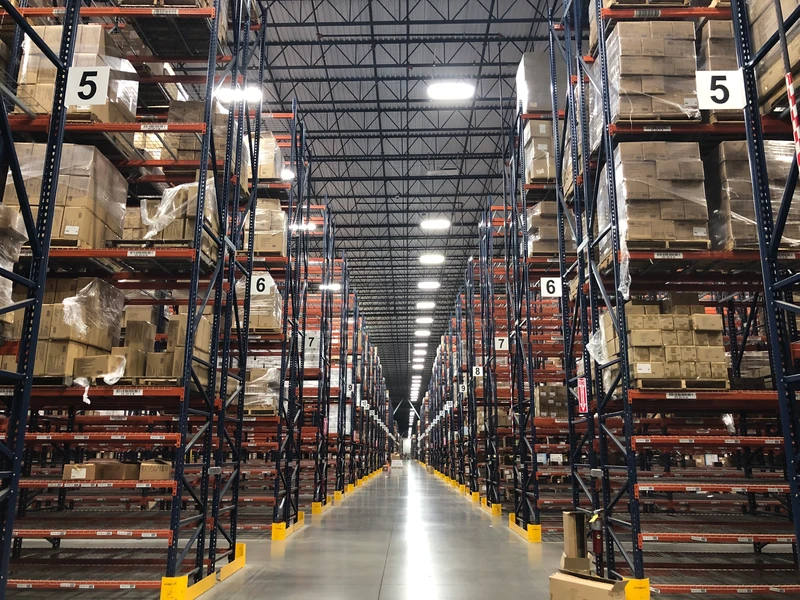 Warehouse interior with high shelves organized in rows, filled with fulfillment boxes and packages. Numbered signs mark the aisles. A wide, well-lit aisle runs down the center.