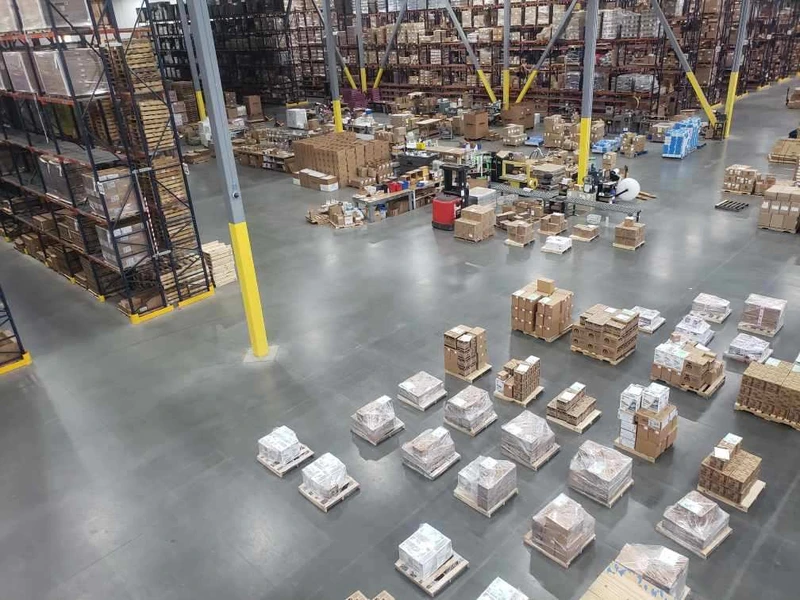 A spacious warehouse with tall shelving units filled with boxes. Several stacks of boxed goods are arranged on the floor, reflecting an efficient logistics system. Workers and machinery are visible in the background, busy picking items for shipment.