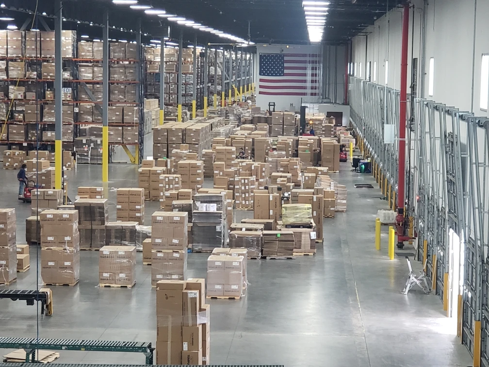 A large warehouse filled with stacks of boxes and pallets. An American flag hangs on the back wall. Forklifts and workers are visible, busy with fulfillment tasks and moving items around the shipping facility.