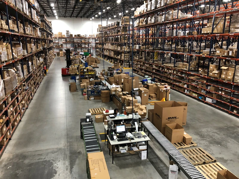 A large fulfillment warehouse filled with rows of tall shelves stocked with various boxes and goods. In the middle, workers are managing packages on a conveyor belt and workstations.